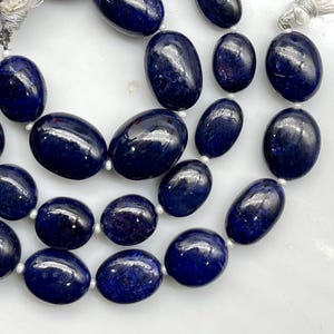May include: A strand of polished, deep blue oval-shaped beads arranged in a multi-row formation. The smooth, dark blue stones feature subtle natural variations and are separated by small white spacers, finished with a light-colored tassel at the end.