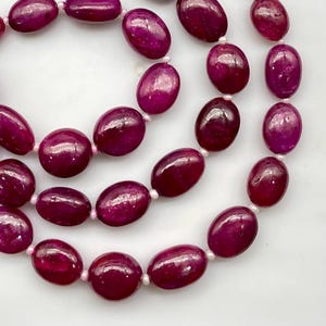 May include: A strand of polished, oval-shaped magenta beads arranged in a circular pattern. Each smooth, deep pink stone is separated by a tiny, round, white spacer bead, creating a continuous, uniform sequence of vibrant, glossy, dark red-purple elements.