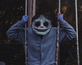 May include: A person wearing a grey sweater and a white mask with a wide smile and red eyes is sitting on a swing set in a dark forest.