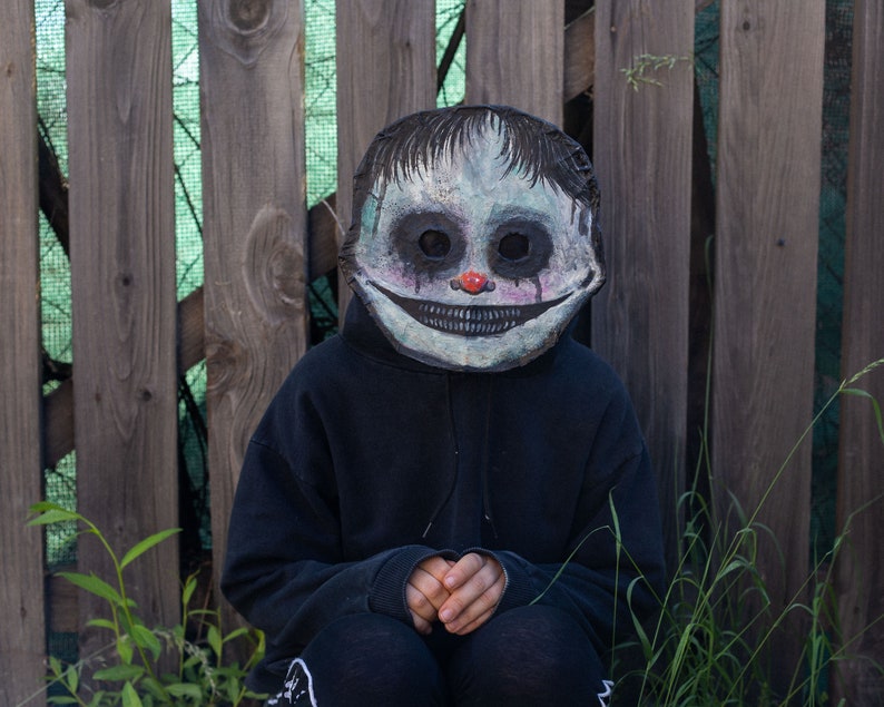 May include: A person wearing a black hoodie and a creepy, hand-painted mask with a wide smile, red nose, and black eyes. The mask is made of paper mache and has a distressed look. The person is sitting in front of a wooden fence.