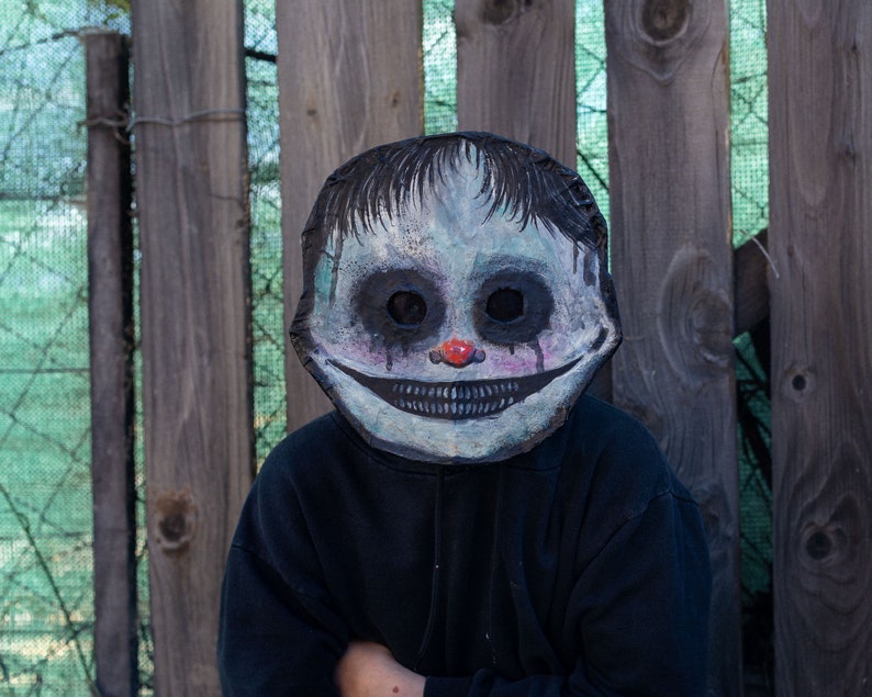 May include: A person wearing a creepy, white and blue painted mask with a wide, toothy grin and red nose. The mask has black eyes and appears to be made of paper mache. The person is wearing a black hooded sweatshirt.