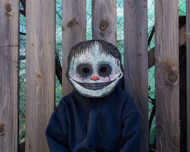May include: A person wearing a black hoodie with a creepy, hand-painted mask. The mask is white with black eyes, a red nose, and a wide, toothy grin.