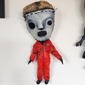 May include: A handmade doll with a mask and red jumpsuit. The mask is painted grey with black eye holes and a large open mouth. The doll has short, light brown hair and is wearing a red jumpsuit with black shoes and gloves. The doll is mounted on a white wall.