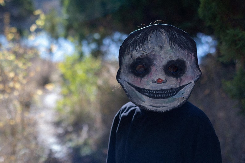 May include: A person wearing a white and black creepy mask with a wide smile and red nose. The mask has black eyes and dark streaks of paint running down the cheeks. The person is wearing a black hooded sweatshirt.