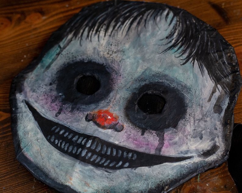 May include: A creepy, hand-painted paper mache mask with a wide, toothy grin, black eyes, and a red nose. The mask is painted in shades of blue, white, and black, with hints of pink and purple.
