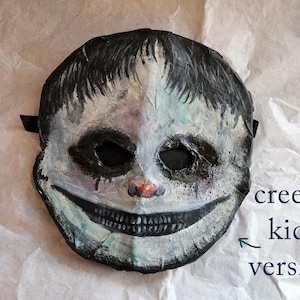 May include: A creepy, white and black painted mask of a child with a wide, toothy grin. The mask has a black strap and is made of paper mache.