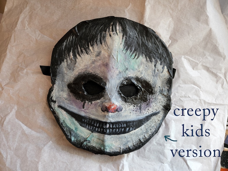 May include: A creepy, white and black painted mask of a child with a wide, toothy grin. The mask has a black strap and is made of paper mache.