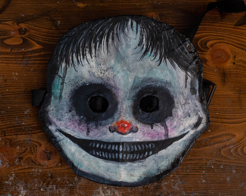May include: A creepy, hand-painted mask with a wide, toothy grin, black eyes, and a red nose. The mask is made of paper mache and has a distressed, aged look.