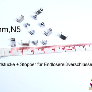 Könnte beinhalten: Nahaufnahme von silbernen Metall-Reißverschluss-Schieber-Stoppern und Endstücken, die 6 mm groß sind. Die Stopper sind in einem dreieckigen Muster auf einer weißen Oberfläche angeordnet, wobei ein weißes Maßband horizontal über den unteren Rand des Bildes verläuft. Der Text "6mm,N5" ist in der oberen linken Ecke des Bildes sichtbar. Der Text "Endstücke + Stopper für Endlosereißverschlüsse" ist am unteren Rand des Bildes sichtbar.