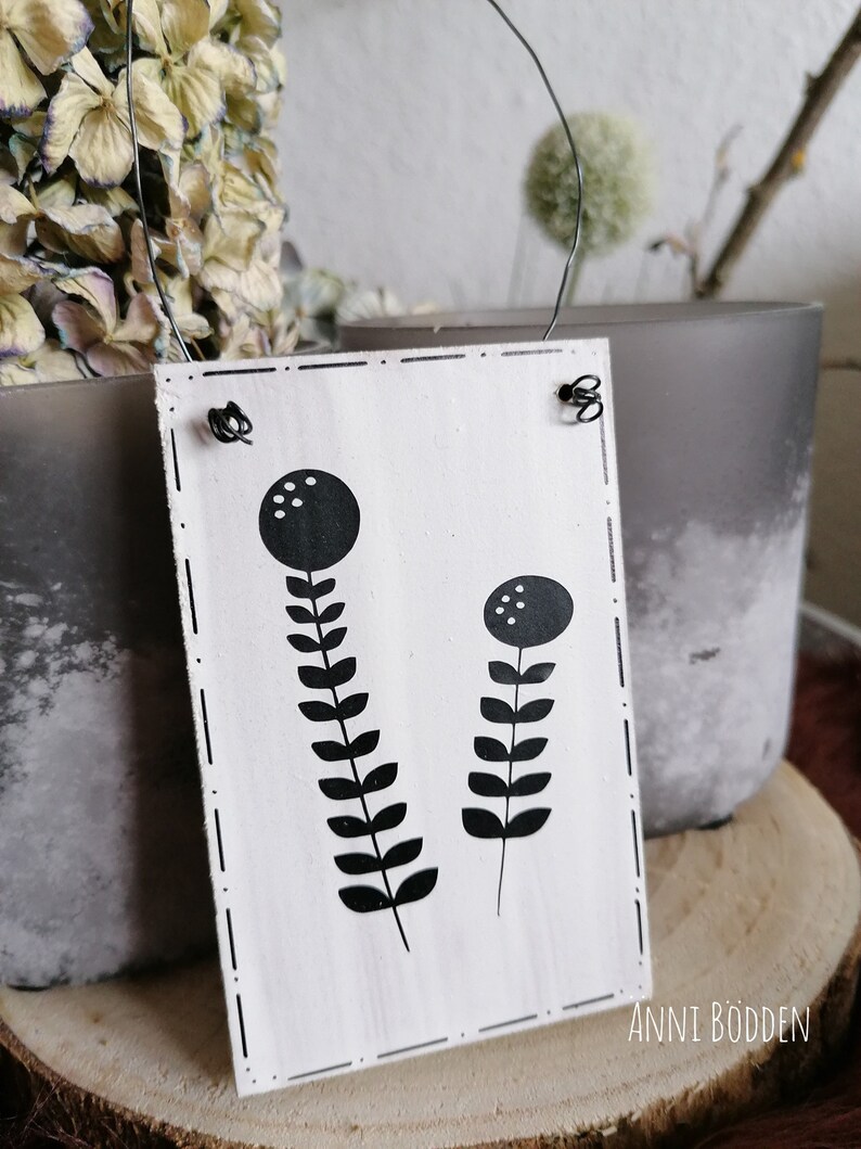 May include: A rectangular white wooden sign with black floral illustrations and the text "ANNI B&Ouml;DDEN". The sign has a wire hanger and is decorated with black dots and dashes. The background includes dried flowers and a gray vase.
