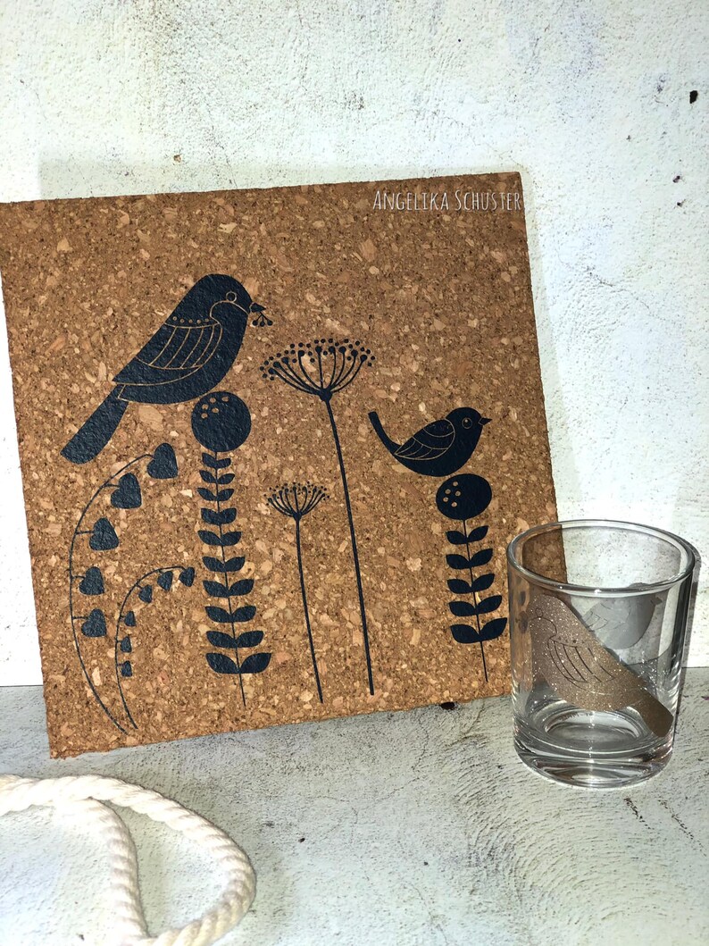 May include: A cork coaster with a black bird and floral design, and a clear glass votive holder with a bird design. The coaster has the text "ANGELIKA SCHUSTER". The design features stylized birds and plants.