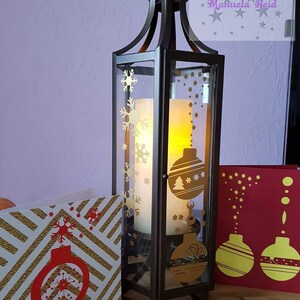 Puede incluir: Una linterna de metal oscuro con paneles de vidrio, adornada con dise&ntilde;os dorados de copos de nieve y adornos. En el interior, se ve una vela encendida. Tarjetas navide&ntilde;as con dise&ntilde;os dorados y rojos se colocan cerca.