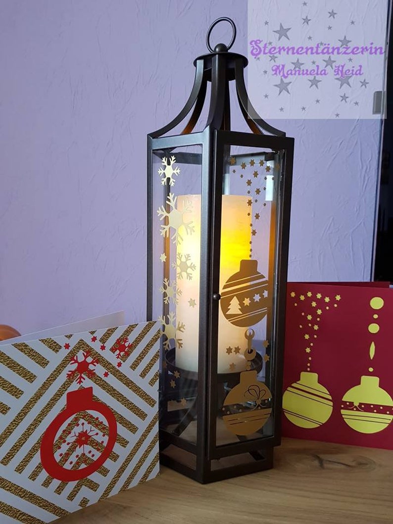 Puede incluir: Una linterna de metal oscuro con paneles de vidrio, adornada con dise&ntilde;os dorados de copos de nieve y adornos. En el interior, se ve una vela encendida. Tarjetas navide&ntilde;as con dise&ntilde;os dorados y rojos se colocan cerca.