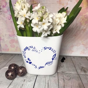 Może przedstawiać: Białe kwiaty hiacyntu w białym ceramicznym wazonie z niebieskim wzorem serca. Wazon stoi na drewnianej powierzchni z trzema ozdobnymi kulami w kolorze miedzi.