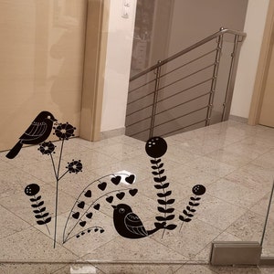 May include: Black decorative bird and floral decals applied to a glass door. The decals include stylized birds perched on branches with flowers and heart-shaped leaves. The door is in a hallway with a staircase.