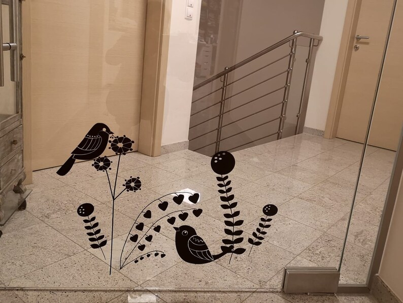 May include: Black decorative bird and floral decals applied to a glass door. The decals include stylized birds perched on branches with flowers and heart-shaped leaves. The door is in a hallway with a staircase.