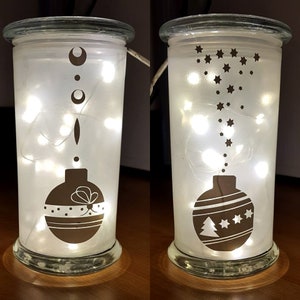Puede incluir: Dos faroles de cristal esmerilado con luces de cadena en el interior. Un tarro tiene un dise&ntilde;o de adorno marr&oacute;n con un lazo y lunares. El otro tarro tiene un dise&ntilde;o de adorno marr&oacute;n con un &aacute;rbol de Navidad y estrellas.