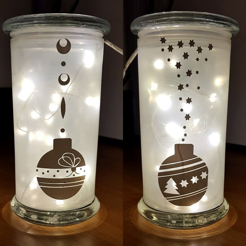 Puede incluir: Dos faroles de cristal esmerilado con luces de cadena en el interior. Un tarro tiene un dise&ntilde;o de adorno marr&oacute;n con un lazo y lunares. El otro tarro tiene un dise&ntilde;o de adorno marr&oacute;n con un &aacute;rbol de Navidad y estrellas.