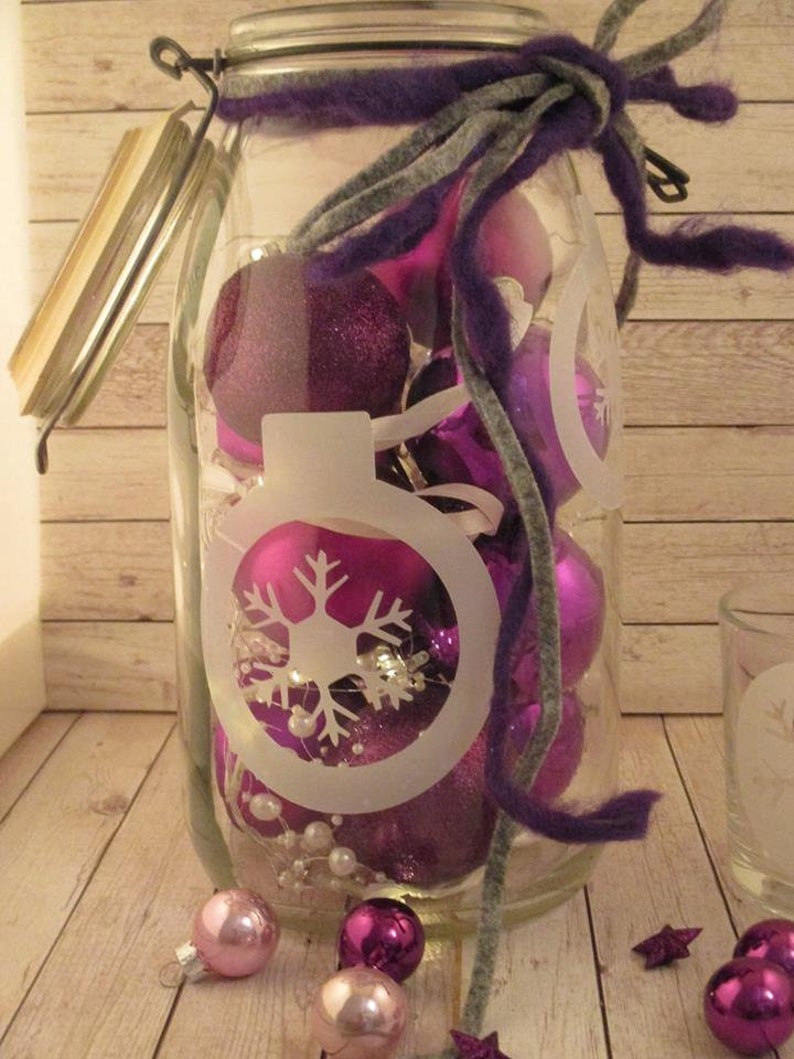 Puede incluir: Un gran tarro de cristal lleno de adornos navide&ntilde;os morados y rosas. El tarro tiene un dise&ntilde;o de copo de nieve blanco y est&aacute; atado con fieltro morado y gris. Varios adornos y decoraciones en forma de estrella est&aacute;n esparcidos en la base.