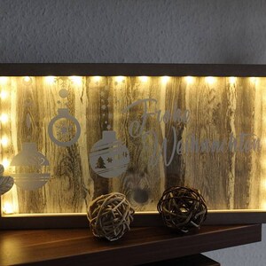Puede incluir: Decoraci&oacute;n navide&ntilde;a rectangular de madera con marco iluminado. El panel de madera tiene un patr&oacute;n de vetas y presenta adornos navide&ntilde;os grabados y las palabras "Frohe Weihnachten". Dos esferas decorativas est&aacute;n al frente.