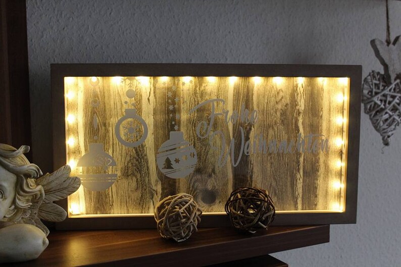 Puede incluir: Decoraci&oacute;n navide&ntilde;a rectangular de madera con marco iluminado. El panel de madera tiene un patr&oacute;n de vetas y presenta adornos navide&ntilde;os grabados y las palabras "Frohe Weihnachten". Dos esferas decorativas est&aacute;n al frente.