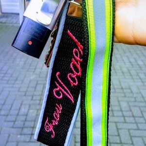 Ein schwarzer und grüner Schlüsselanhänger mit einem weißen reflektierenden Streifen. Der Schlüsselanhänger hat ein schwarzes Stoff-Etikett mit dem rosa gestickten Text "Frau Vogel!"
