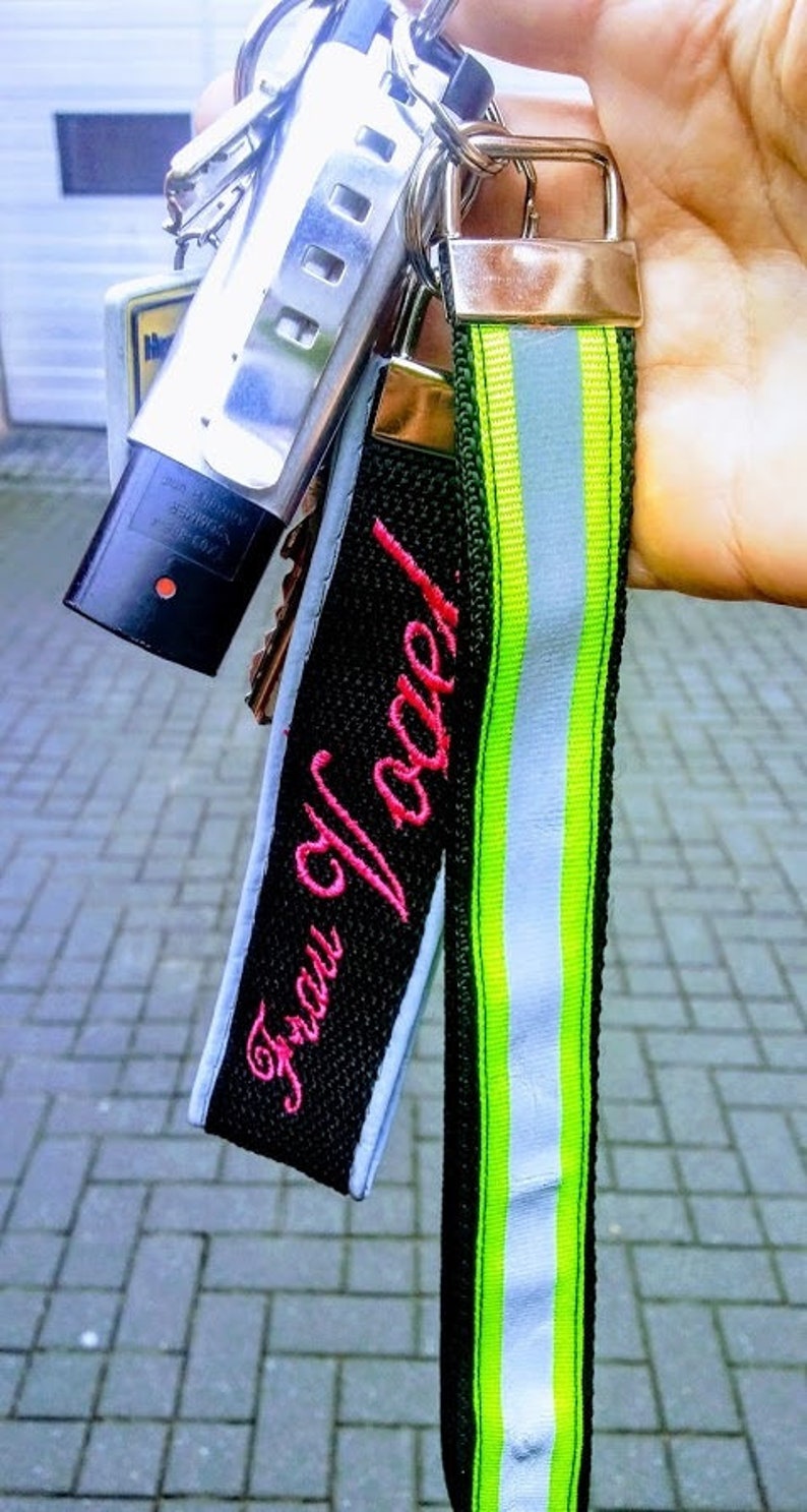 Ein schwarzer und grüner Schlüsselanhänger mit einem weißen reflektierenden Streifen. Der Schlüsselanhänger hat ein schwarzes Stoff-Etikett mit dem rosa gestickten Text "Frau Vogel!"