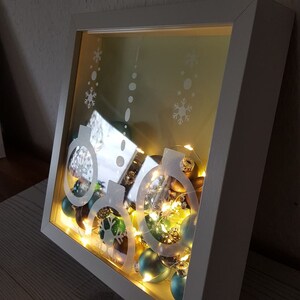 Puede incluir: Una caja de sombra con marco blanco con un tema navide&ntilde;o festivo. La caja contiene adornos de color verde azulado y marr&oacute;n, iluminados por luces de hadas blancas c&aacute;lidas. El vidrio est&aacute; grabado con dise&ntilde;os de copos de nieve y adornos.
