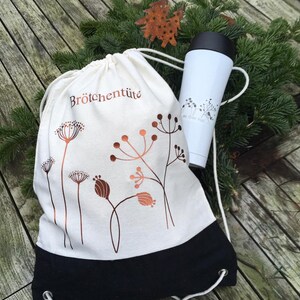 May include: A white drawstring bag with the text "Br&ouml;tchent&uuml;te" and copper-colored floral designs, paired with a white travel mug featuring a black lid and floral graphics. The bag has a black bottom.