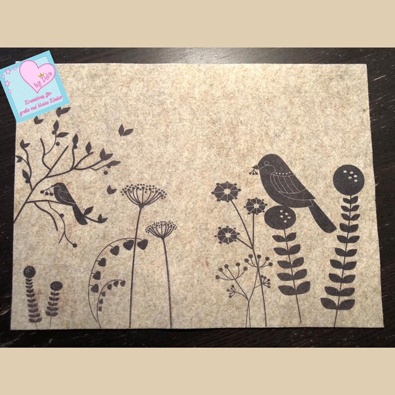 May include: A beige felt placemat with a black floral and bird design. The design includes stylized flowers, leaves, and two birds perched on branches. A small tag in the upper left corner has text and a pink heart.
