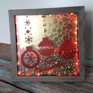 Puede incluir: Decoraci&oacute;n navide&ntilde;a enmarcada con un marco de madera gris. La obra presenta adornos rojos, copos de nieve y el texto "Happy Winter". El fondo es dorado con peque&ntilde;as cuentas doradas y luces rojas.