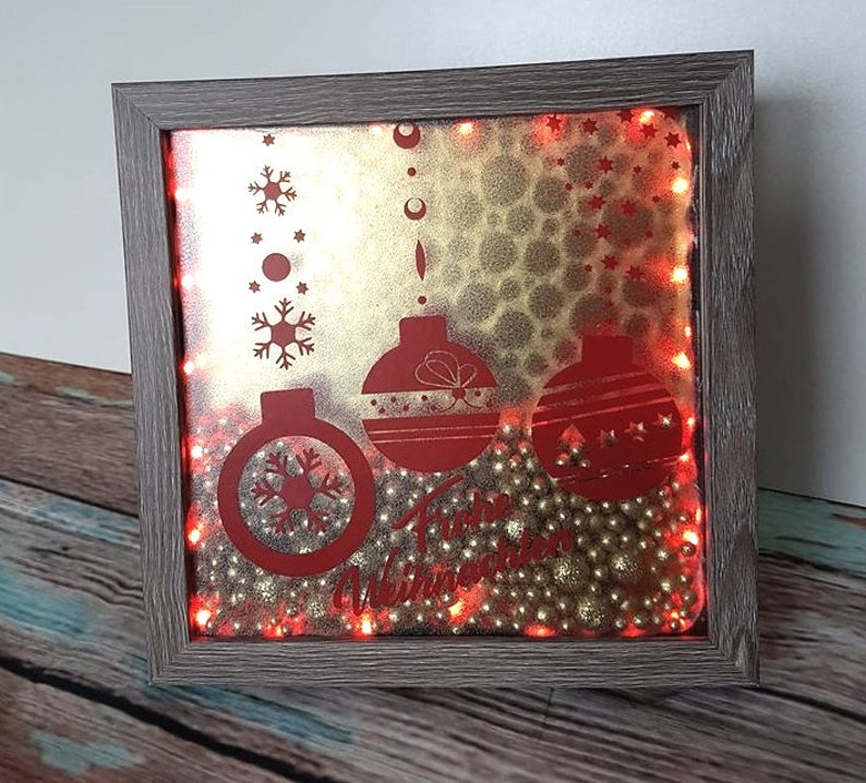 Puede incluir: Decoraci&oacute;n navide&ntilde;a enmarcada con un marco de madera gris. La obra presenta adornos rojos, copos de nieve y el texto "Happy Winter". El fondo es dorado con peque&ntilde;as cuentas doradas y luces rojas.