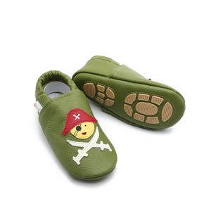 Op de afbeelding: Een paar groene leren babyschoentjes met een piratengezicht en een ontwerp met gekruiste zwaarden. De schoenen hebben een bruine rubberen zool met de merknaam "Liya's" op de onderkant.