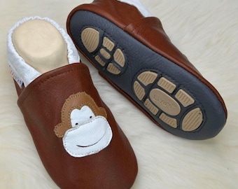 May include: A pair of brown leather soft-sole slippers featuring a white monkey face appliqué on the front. The slippers have a white elasticated collar and a dark grey sole with tan rubber traction pads embossed with the brand name Liya's.