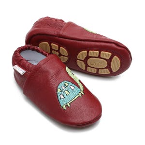 Op de afbeelding: Een paar rode leren babyschoentjes met een geborduurd schildpadontwerp in groen en blauw. De schoenen hebben een textuurzool en zeggen "Liya's" op de onderkant.