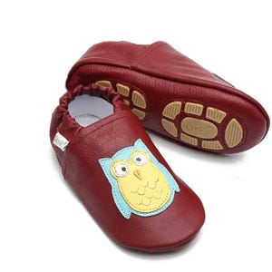 Op de afbeelding: Een paar rode leren babyschoentjes met een uil-ontwerp. De schoentjes hebben een zachte, flexibele zool en zijn perfect om te kruipen en te lopen. Het uil-ontwerp is geborduurd op de voorkant van de schoentjes en heeft een blauw lichaam, een gele snavel en zwarte ogen.