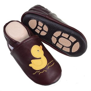 Op de afbeelding: Een paar bruine leren babyschoentjes met een gele eend-applicatie. De schoentjes hebben een zwarte rubberen zool met de tekst "Liya's" op de onderkant.