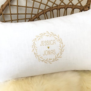 K&ouml;nnte beinhalten: Ein wei&szlig;es Leinenkissen mit einem goldenen gestickten Kranzdesign. Der Kranz enth&auml;lt die Namen "Jessica" und "Jonas" mit einem kleinen Herzen dazwischen.