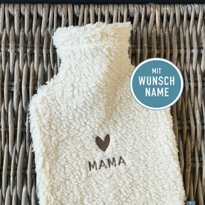 Könnte beinhalten: Cremefarbener Wärmflaschenbezug mit dem Wort "MAMA" und einem braunen Herz. Der Bezug hat eine flauschige Textur und einen runden blauen Aufkleber mit der Aufschrift "MIT WUNSCH NAME".