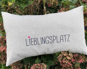Könnte beinhalten: Ein graues Kissen mit dem gestickten Text "LIEBLINGSPLATZ" in Schwarz und einem kleinen pinkfarbenen Herzen über dem Text.
