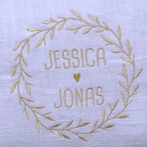 K&ouml;nnte beinhalten: Ein wei&szlig;es Leinenkissen mit einer goldenen gestickten Kranz und den Namen "Jessica" und "Jonas" darin. Ein kleines goldenes Herz befindet sich zwischen den Namen.