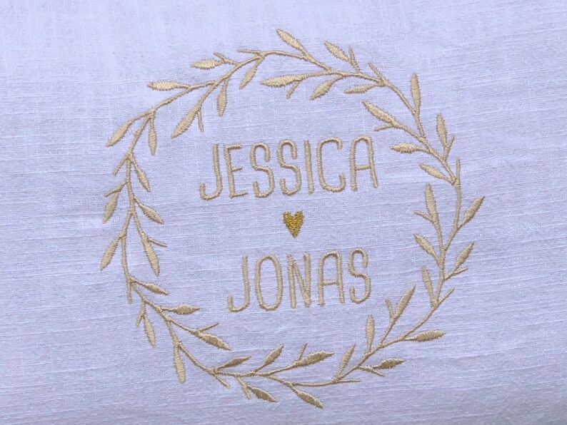 K&ouml;nnte beinhalten: Ein wei&szlig;es Leinenkissen mit einer goldenen gestickten Kranz und den Namen "Jessica" und "Jonas" darin. Ein kleines goldenes Herz befindet sich zwischen den Namen.