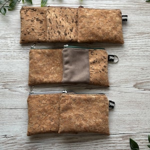 Könnte beinhalten: Drei Kork-Geldbörsen mit Reißverschlüssen werden auf einer hellen Holzoberfläche gezeigt. Die Geldbörsen haben ein natürliches, strukturiertes Aussehen mit Farbvariationen des Korks. Eine Geldbörse hat eine kontrastierende graue Platte. Jede Geldbörse hat ein kleines schwarzes Etikett.