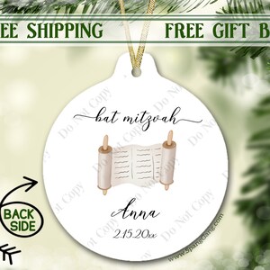 May include: White round bauble with a gold ribbon. The bauble features a watercolour illustration of a Torah scroll with the text "bat mitzvah" and the name "Anna" with the date "2.15.20xx".