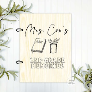 May include: A wooden sign with the text "Mrs. Cox's 2nd Grade Memories" in black lettering. The sign features a black and white illustration of a book with the letters "ABC" on the cover and a pencil holder with pencils.