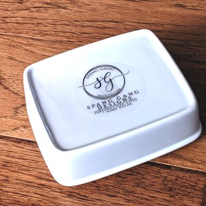 May include: White ceramic square dish with rounded edges. The dish has a sticker on the bottom with the text "Spang Gang Designs.com Personalized Gifts + Home Decor" and a logo with the letters "SG" inside a circle.