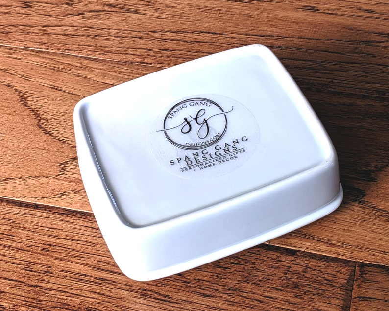May include: White ceramic square dish with rounded edges. The dish has a sticker on the bottom with the text "Spang Gang Designs.com Personalized Gifts + Home Decor" and a logo with the letters "SG" inside a circle.