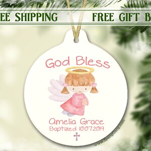 May include: White ceramic ornament with a watercolor illustration of a girl angel with pink wings and a halo. The text "God Bless" is written in pink above the angel and "Amelia Grace Baptized 10.07.2019" is written below the angel.