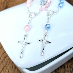 May include: Two silver cross rosaries with pink and blue beads. The rosaries are displayed in a white box. The box has the text "Spang Gang Designs" on it.