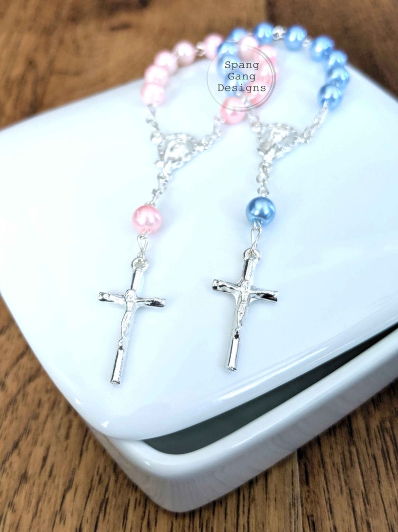 May include: Two silver cross rosaries with pink and blue beads. The rosaries are displayed in a white box. The box has the text "Spang Gang Designs" on it.